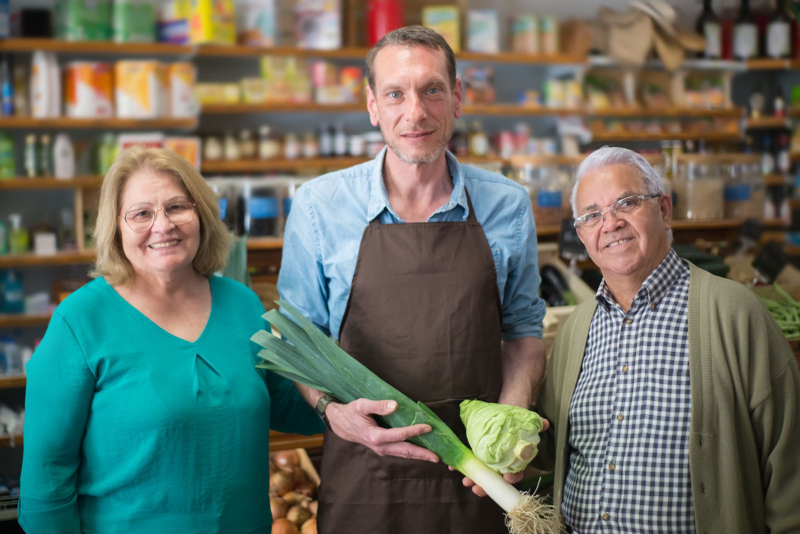 A smiling grocer in an apron with two regular customers holding fresh leeks