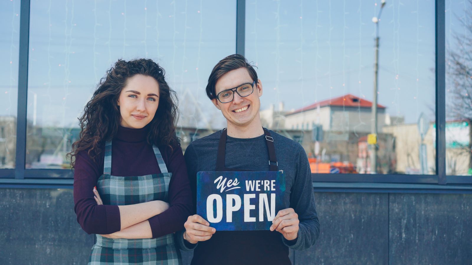 Two smiling shop owners holding a We Are Open sign outside their storefront